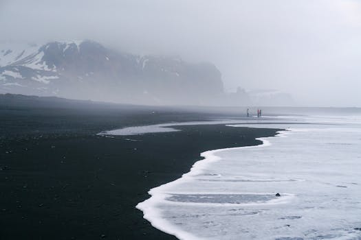 Misty coastal scene with black sand, waves, and distant mountains.