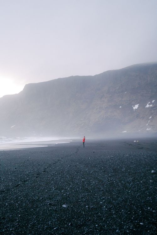 Free A lone person walks on a mist-covered pebbly beach with rugged cliffs in the background. Stock Photo