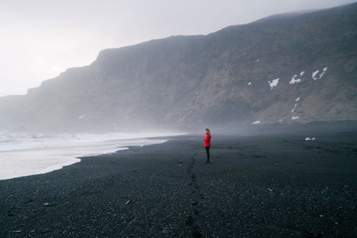 Free A person in a red jacket stands alone on a misty black sand beach under a cliff. Stock Photo