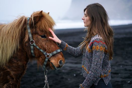 A woman gently pats a horse on a black sand beach, showcasing tranquility by the sea.