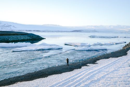 Free A lone person stands on the snowy shore of a serene ice-filled lake in Iceland. Stock Photo