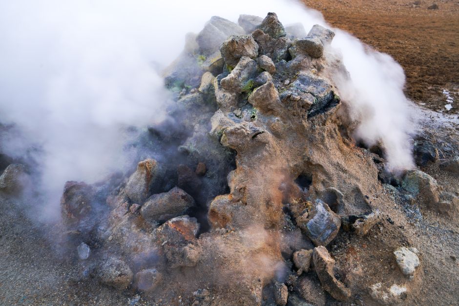 A close-up view of steam emanating from a rocky geothermal formation in a rugged landscape.