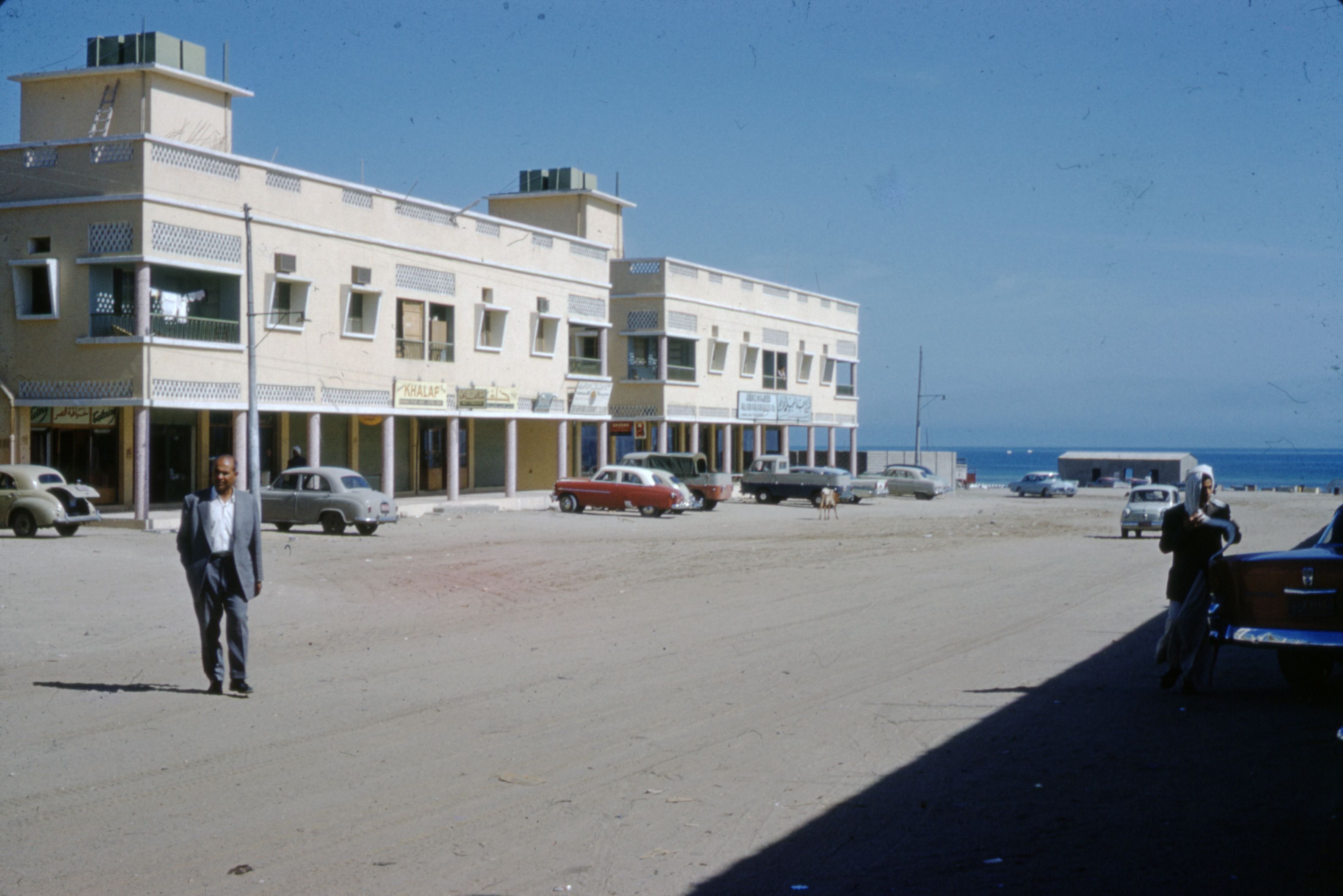 Retro scene of a busy street near the beach with classic cars and people walking.