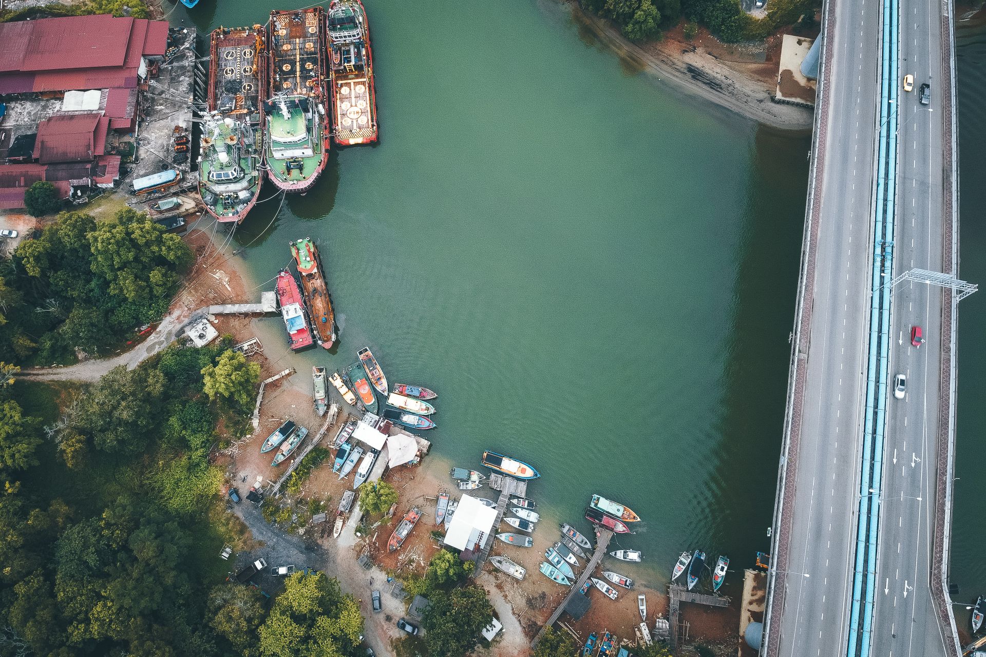 Drone view of cars riding on bridge over channel near various moored boats in tropical countryside