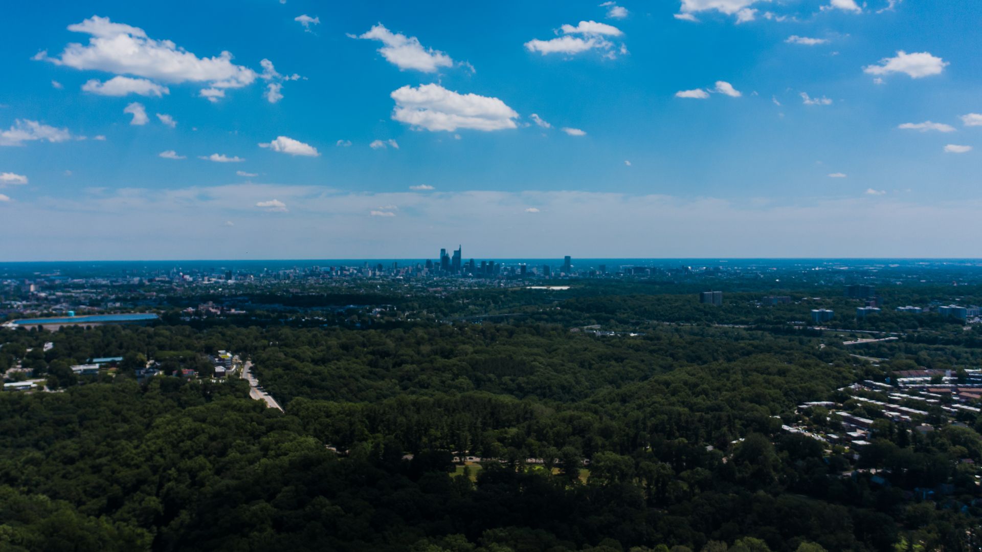 Stunning aerial view of Philadelphia cityscape surrounded by dense greenery under a bright blue sky.