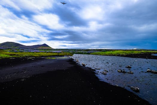 Serene landscape featuring a river, black sand, and mountains under a cloudy sky.