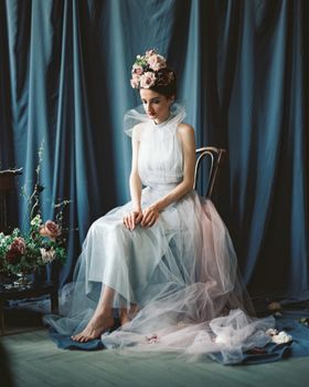 A thoughtful bride in a floral adorned wedding dress sitting indoors, capturing a pensive moment.