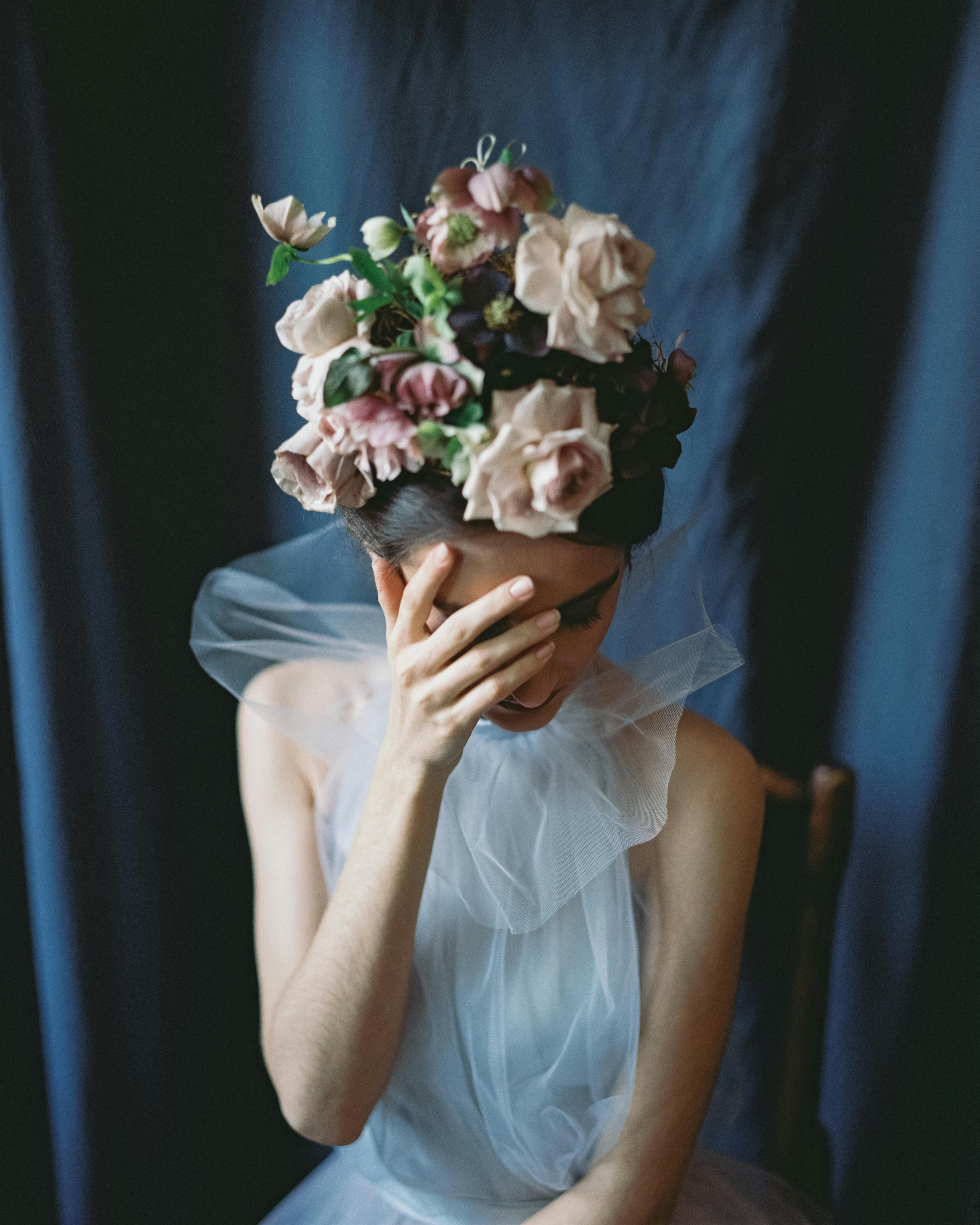 A woman in a stylish dress adorned with a floral headpiece, sitting indoors with a hand on her face.