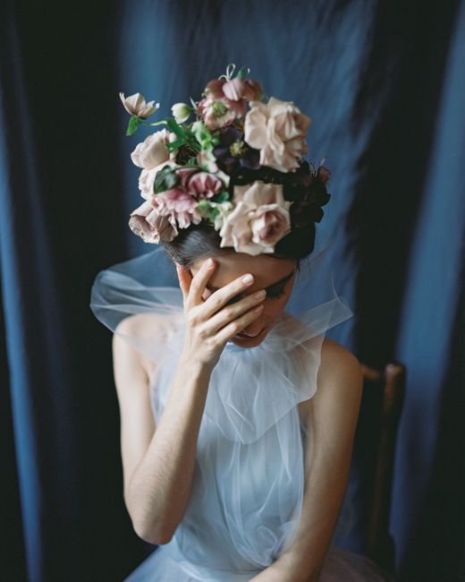 A woman in a stylish dress adorned with a floral headpiece, sitting indoors with a hand on her face.