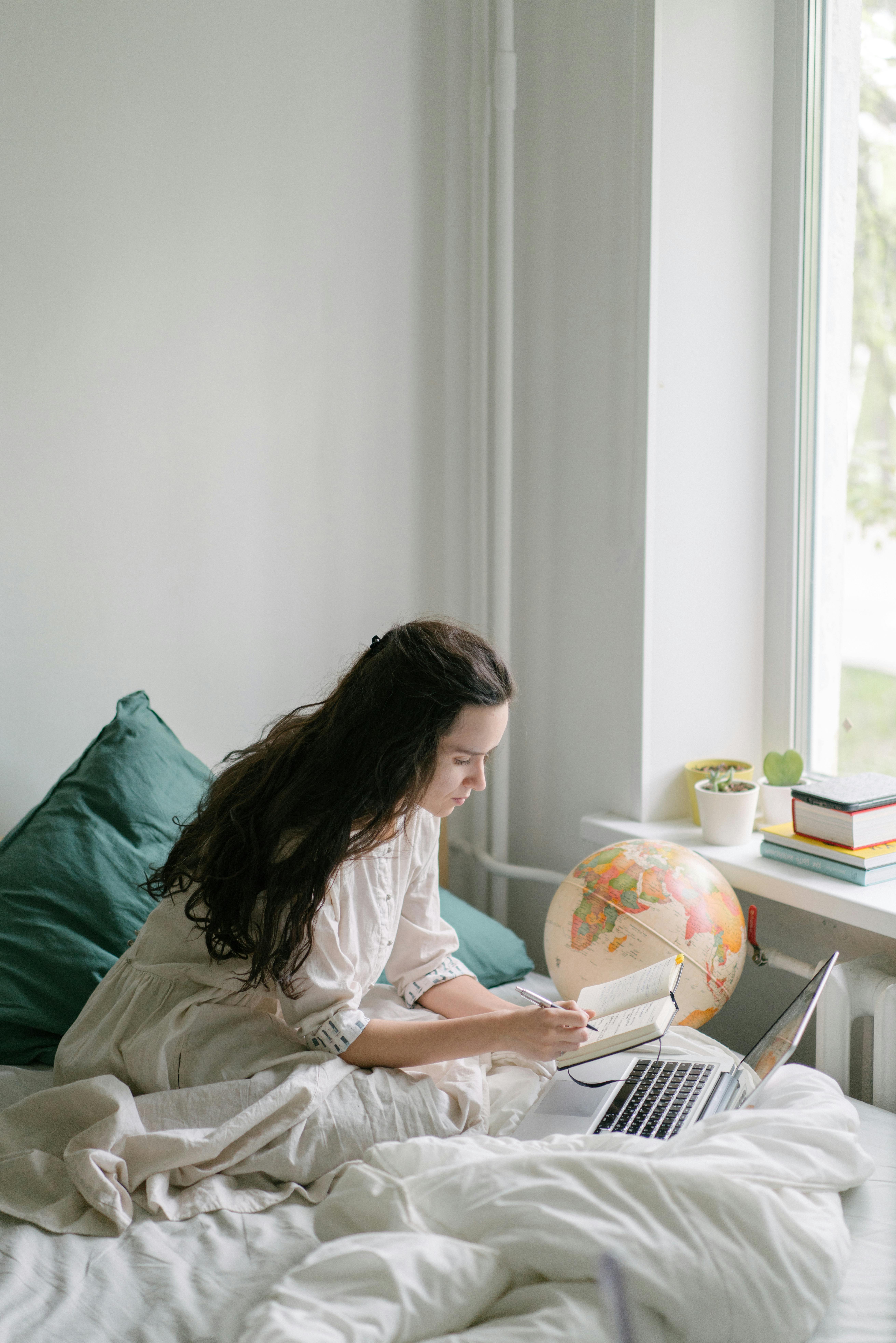 Woman sitting on bed by window, working on a laptop with a notebook. Warm, peaceful atmosphere.