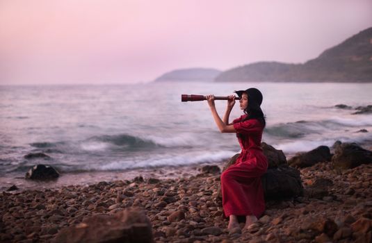 A Woman in Red Dress Sitting on a Rock Holding a Monocular Telescope