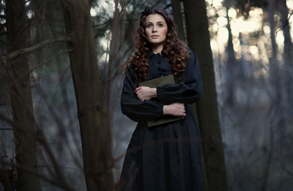 A woman with curly hair holding a book, standing in a dark, mysterious forest.