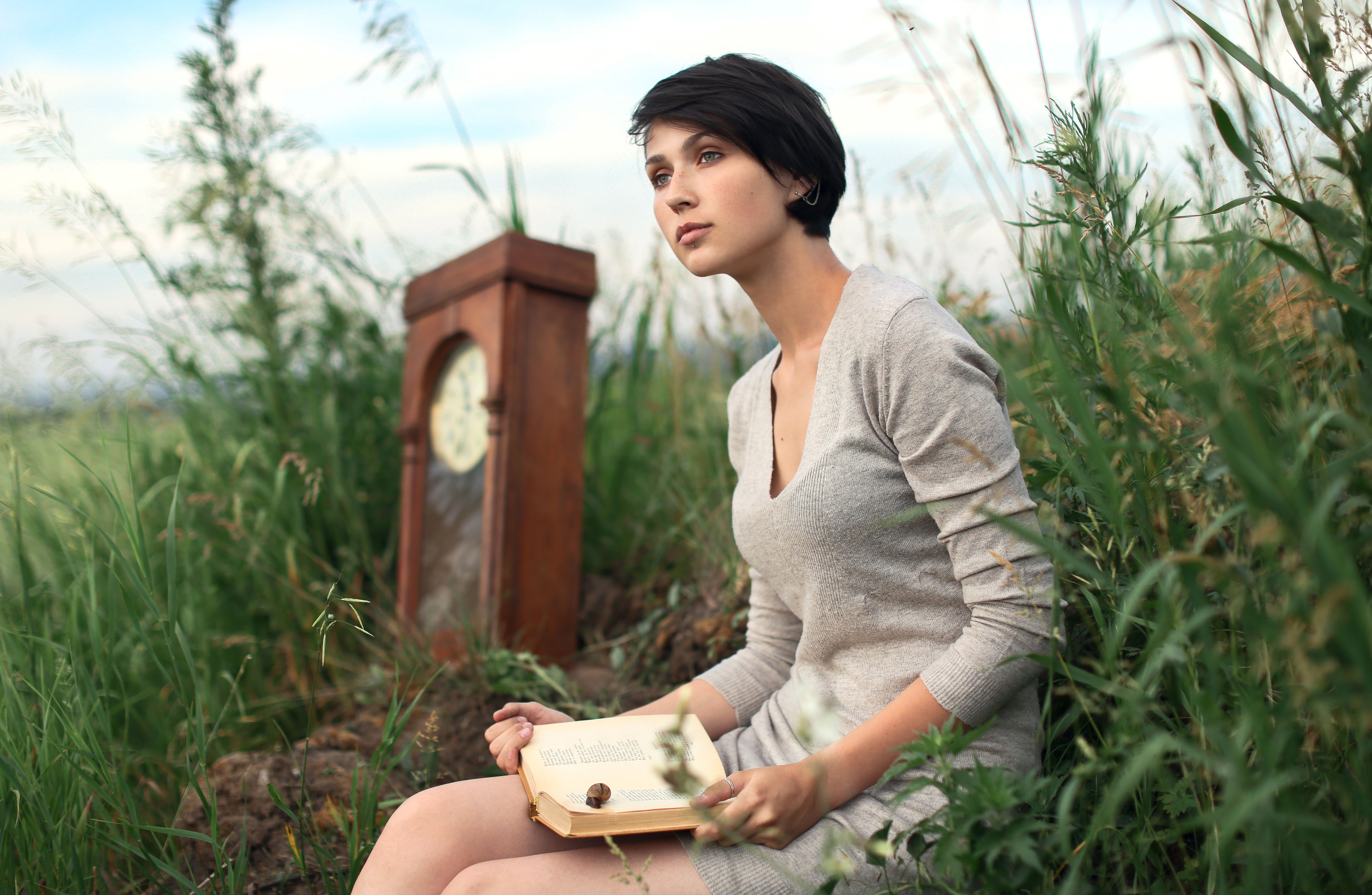A young woman sitting in a field, holding an open book near a vintage clock, surrounded by tall grass.