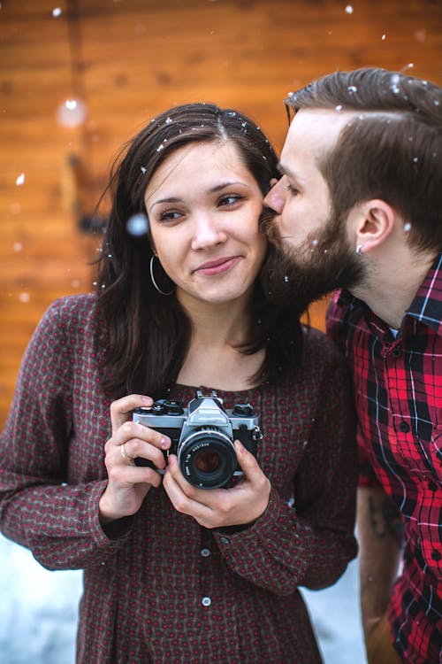Free A Bearded Man Kissing a Woman Holding a Camera Under the Snow Stock Photo