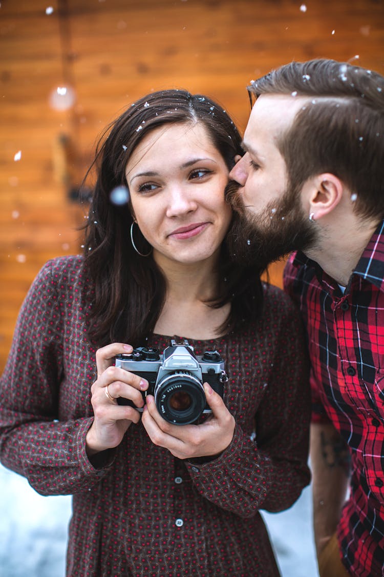 A Bearded Man Kissing A Woman Holding A Camera Under The Snow