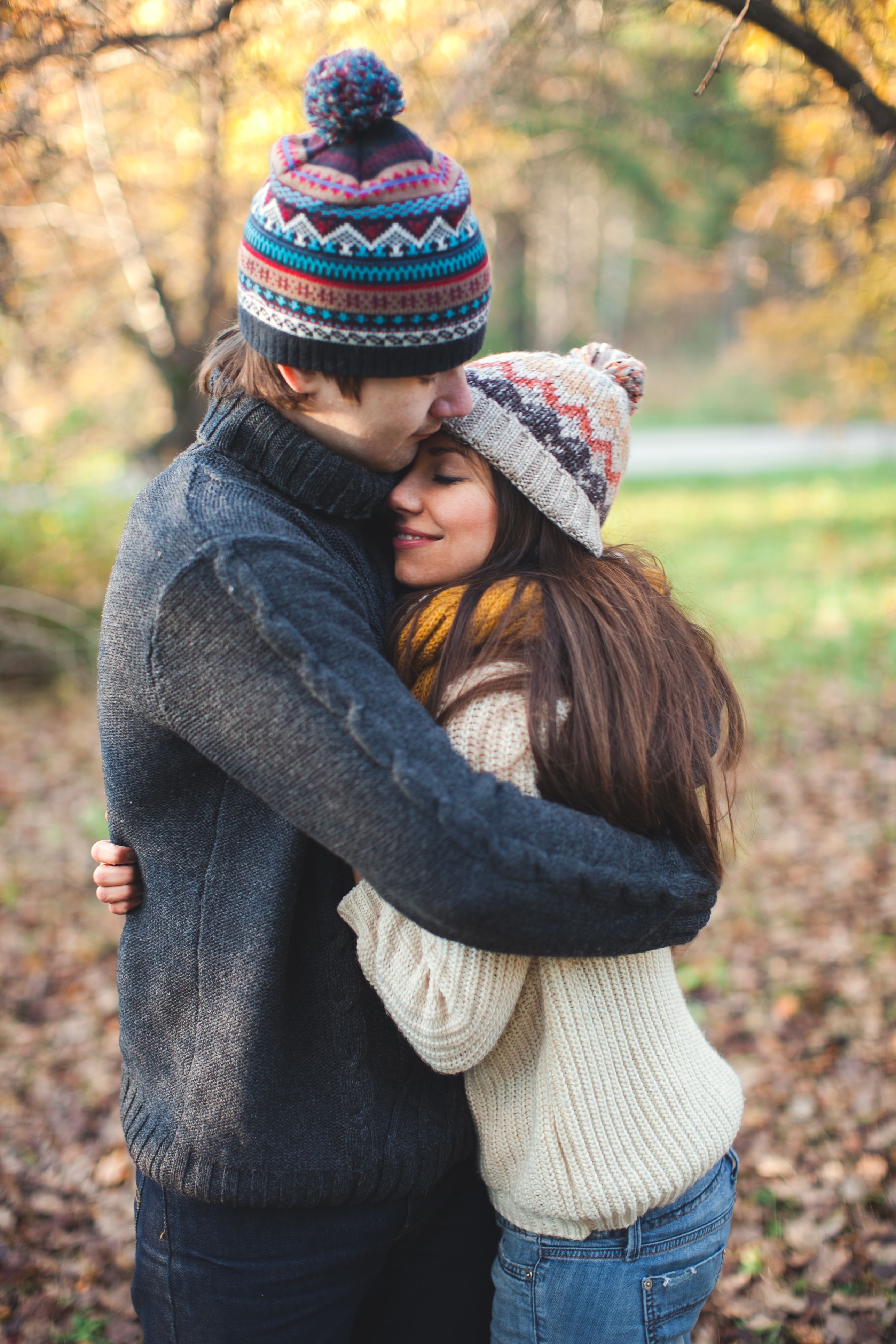 Young couple embracing in a park during fall, wearing cozy knitted hats and sweaters.