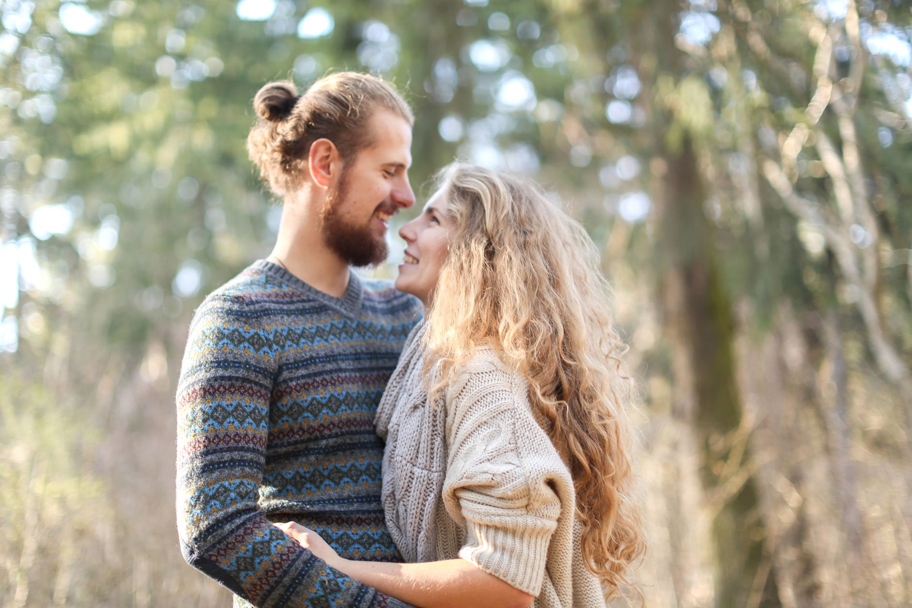 Couple in knitwear sharing affectionate gaze amidst woodland backdrop.