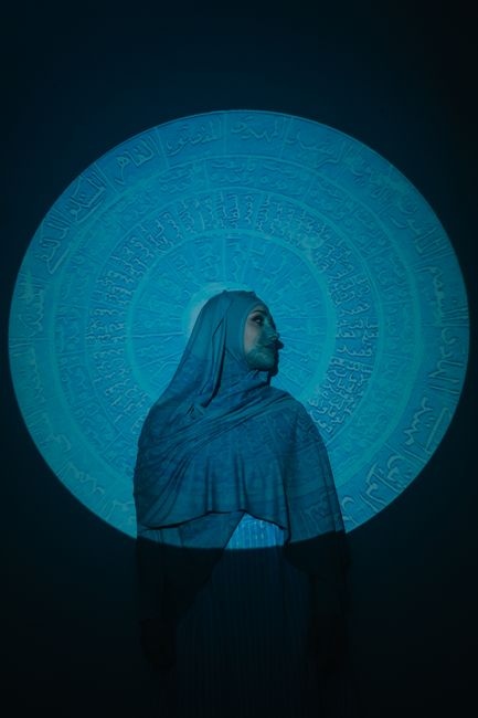 A woman in a hijab stands in front of an artistic blue Arabic calligraphy projection, creating a mysterious aura.