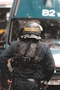 Back view of faceless police officer wearing helmet and protective vest with gun and baton standing in front of police van