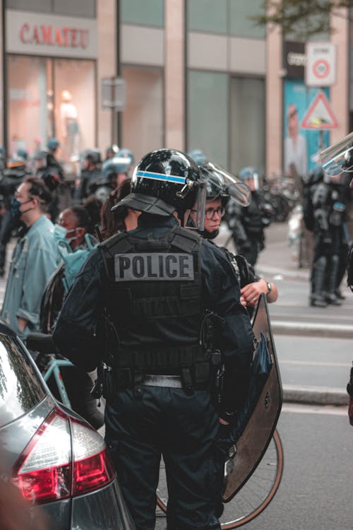 Free Back view of unrecognizable police officer with helmet and shield performing duties on protest while standing near car and protesters in masks Stock Photo