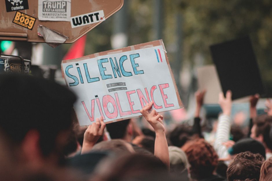 Una multitud se reúne para una protesta pacífica, sosteniendo un cartel que dice 'Silencio = Violencia' en una calle.