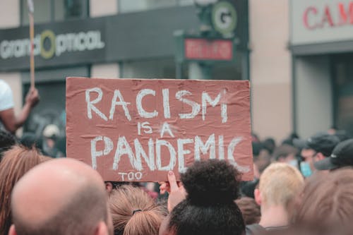 Crowd of people standing and protesting against police atrocity holding handmade sign reading Racism is a pandemic too