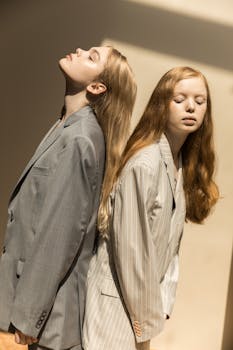 两名穿着时尚西装的女性在室内阳光下背靠背摆姿势拍摄艺术肖像