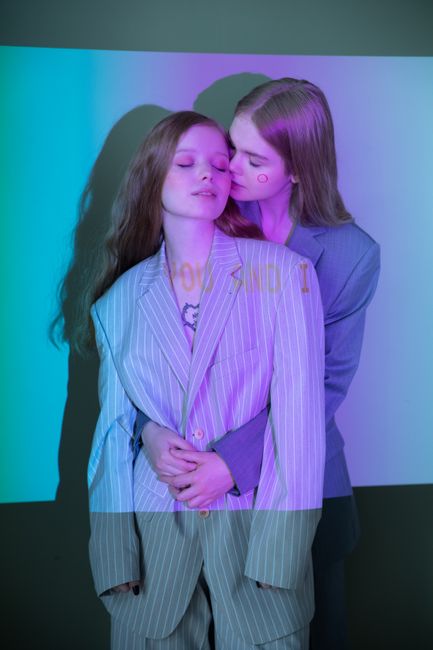 Two women embrace in stylish suits with colorful light projections, symbolizing love