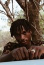 Serious black male with dreadlocks standing and putting hands on metal surface and looking at camera against tree