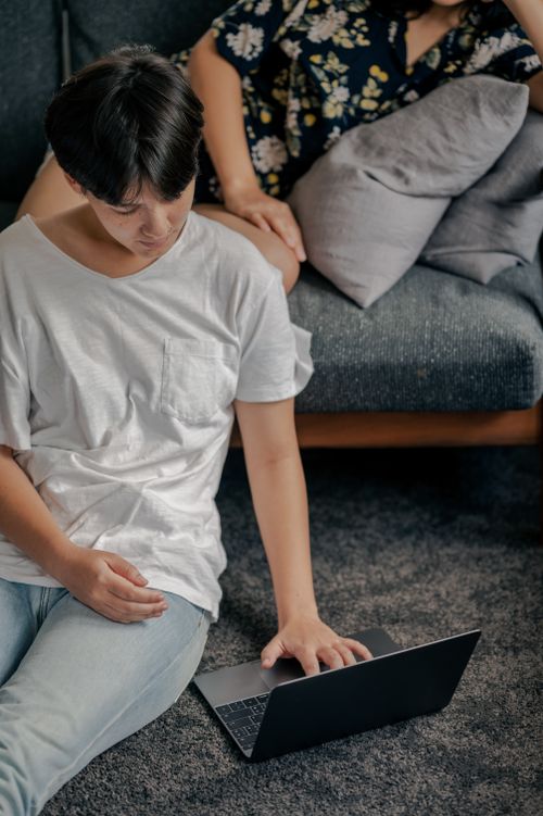 Free A young Asian man sitting on the floor using a laptop in a cozy living room setup with a couch. Stock Photo
