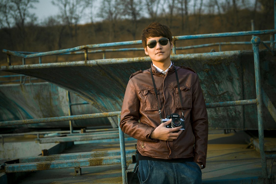 Free Man in Brown Leather Jacket Wearing Black Framed Sunglasses Holding Black Dslr Camera Standing in Front of Black Steel Bar during Daytime Stock Photo