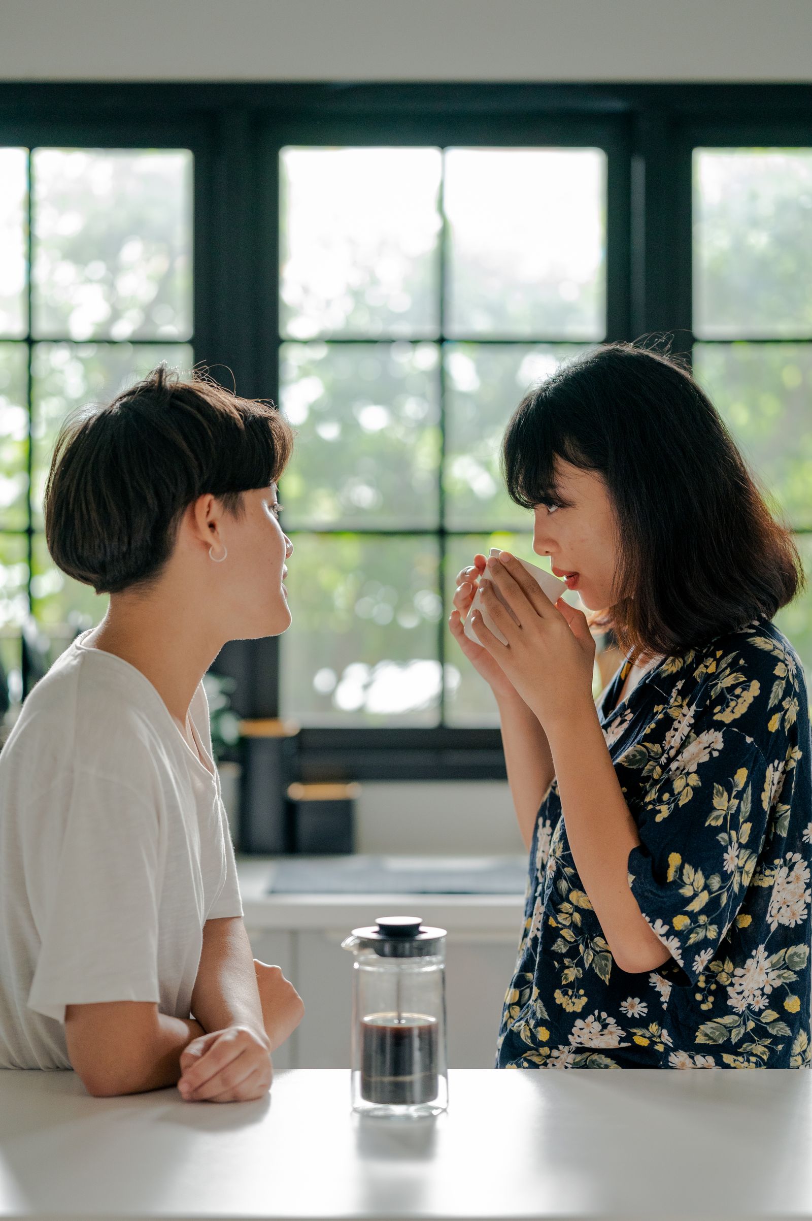 Two people talking over coffee beside a kitchen window