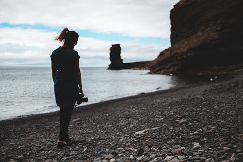 Free Full body of unrecognizable traveler in casual clothing carrying photo camera while having stroll on rocky shore of calm ocean Stock Photo