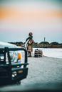 Side view of African female wearing traditional dress standing on sandy shore with bags and car in front
