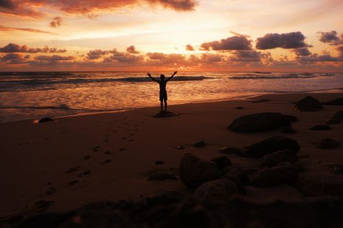 Free A dramatic beach scene featuring a man's silhouette against a vibrant sunset sky and ocean waves. Stock Photo