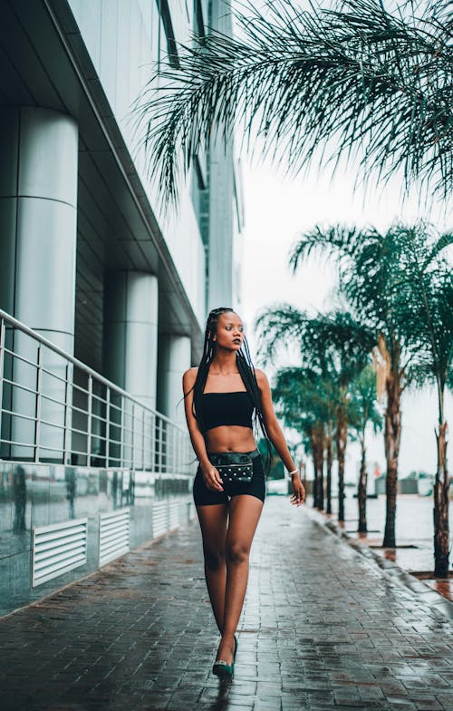 Full body of confident young African American female wearing black top and shorts walking along pavement and looking away