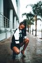 Full body trendy black female with dreadlocks in casual wear and heels squatting on pavement with palm trees behind