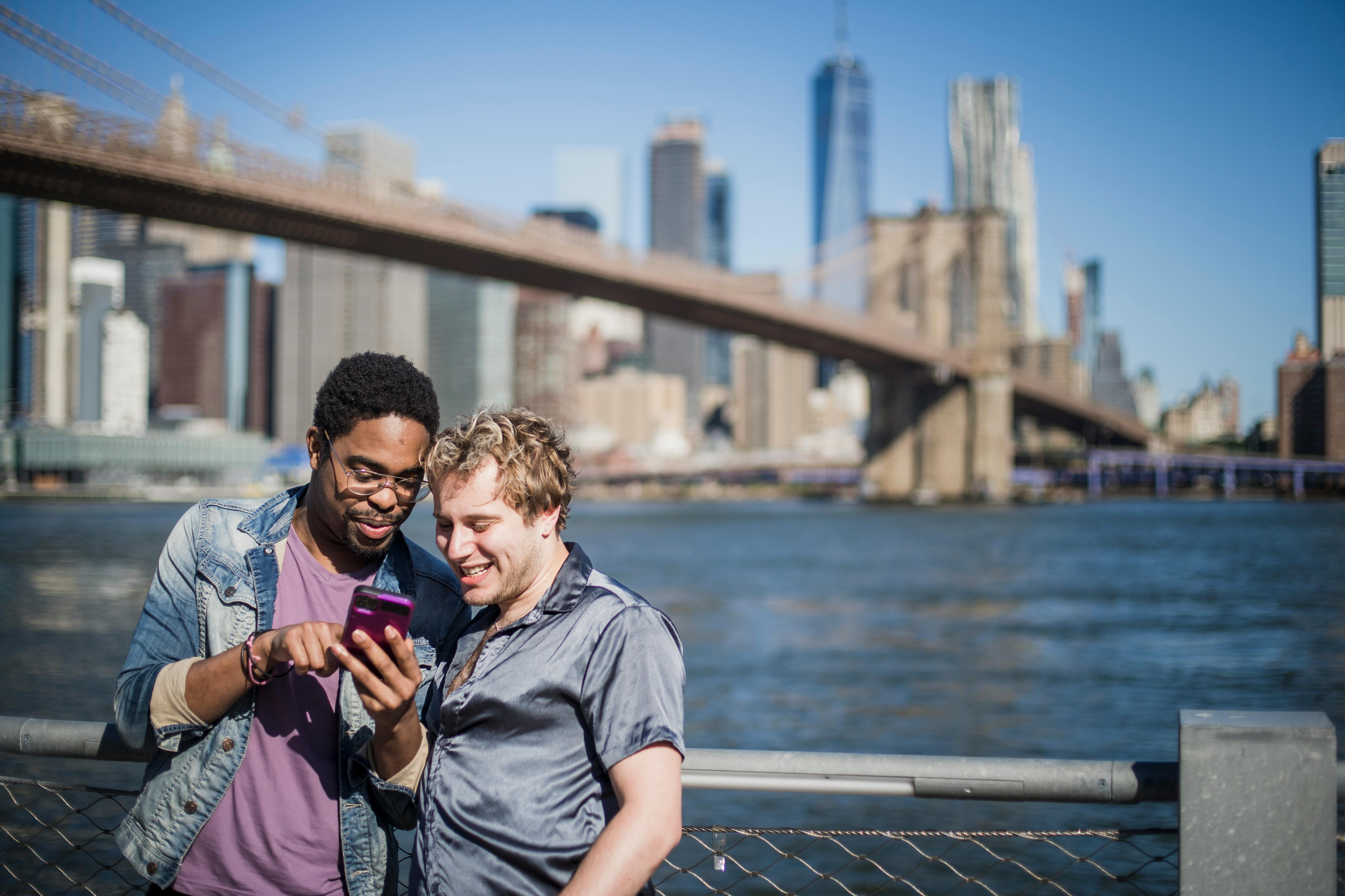 两个朋友在布鲁克林大桥和纽约天际线的背景下，浏览智能手机时微笑着