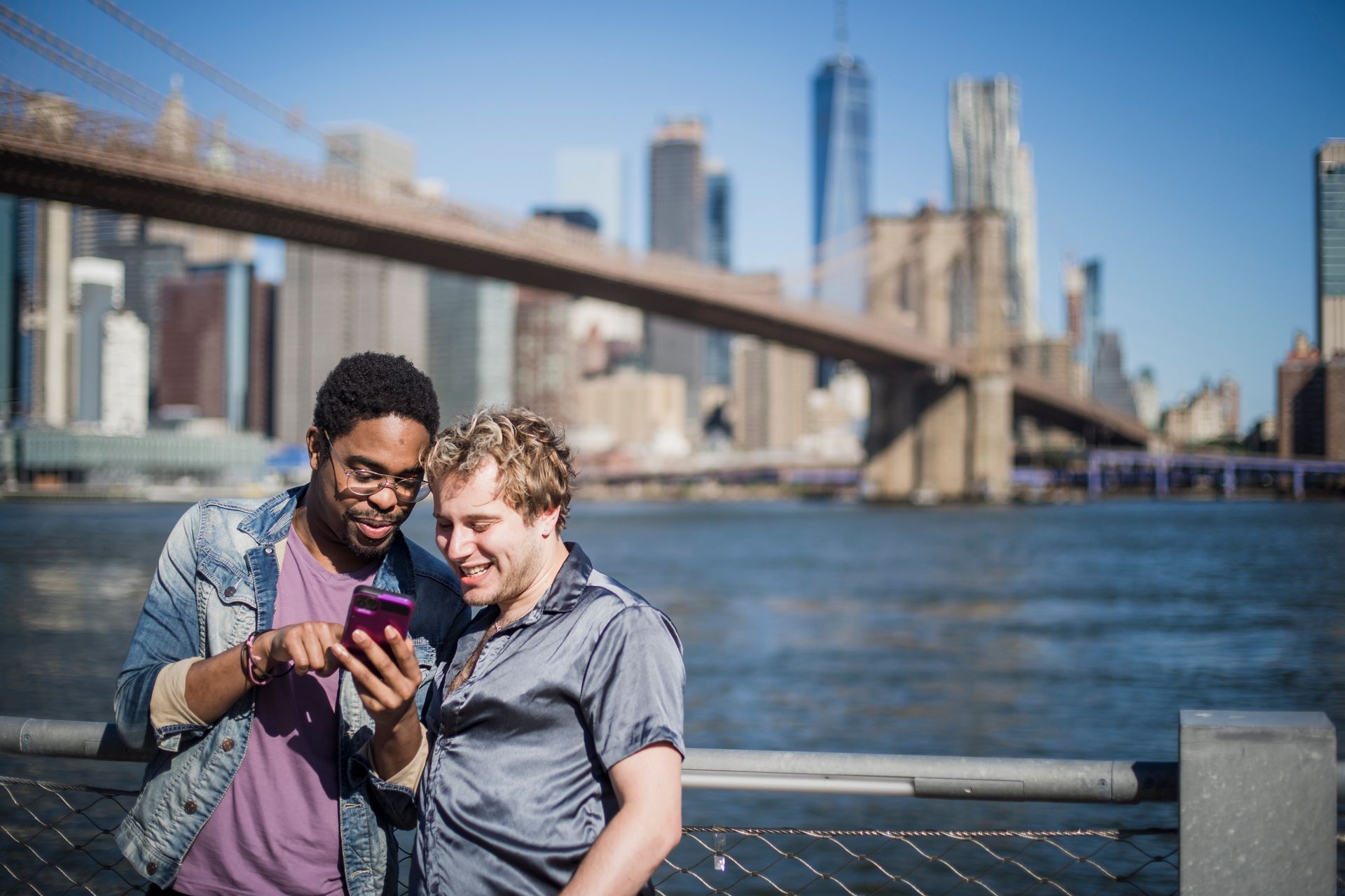 Two friends smiling while browsing a smartphone with the Brooklyn Bridge and NYC skyline in the background.