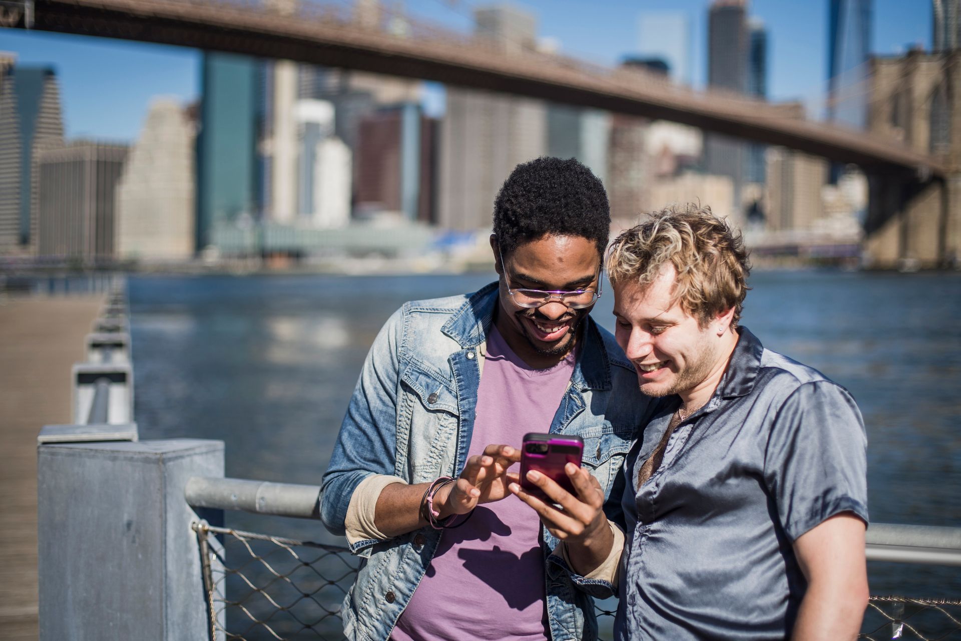 Joyful interracial LGBT couple smiling and looking at smartphone near urban bridge in daylight.