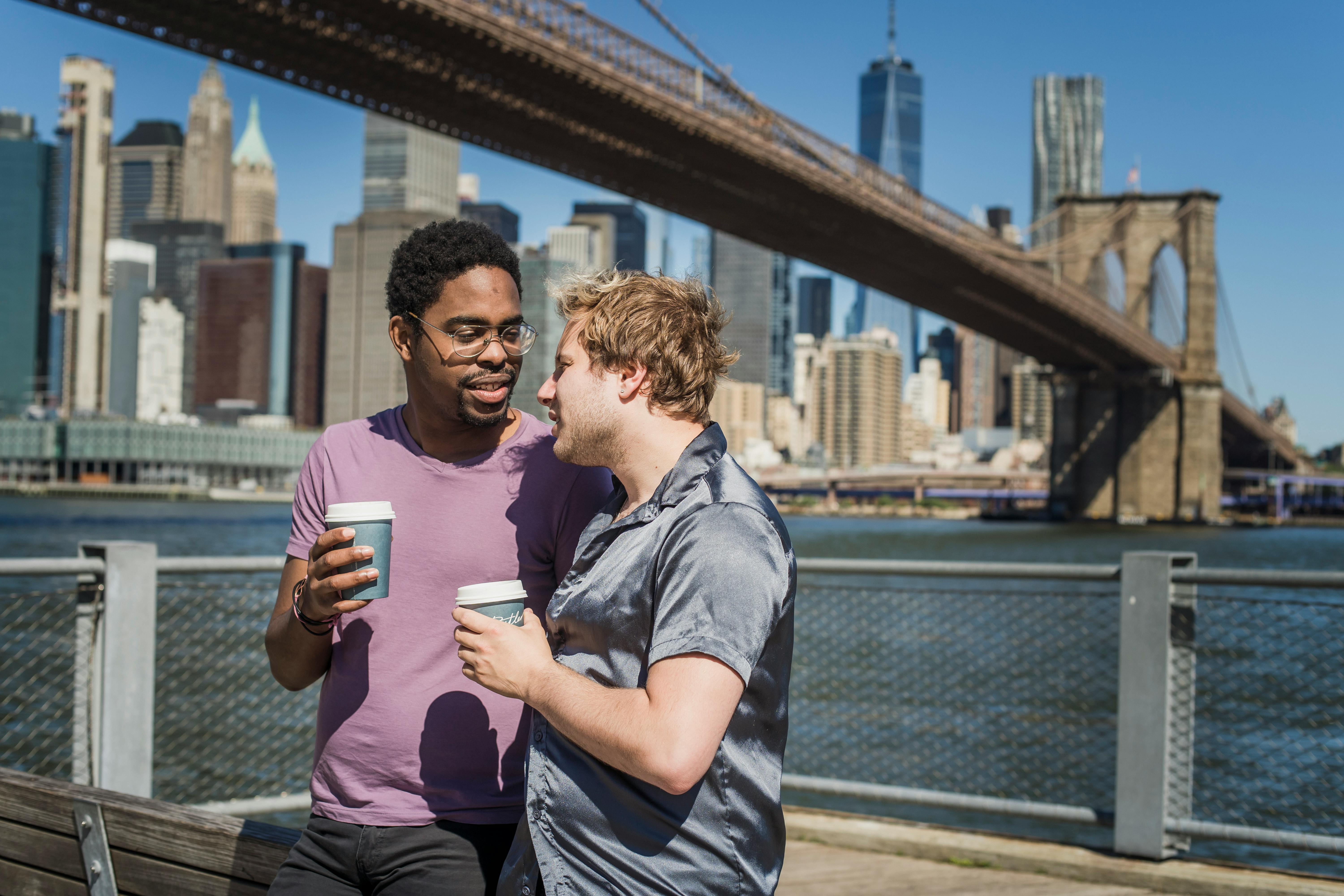 两名男子在户外享用咖啡，背景是纽约市天际线和布鲁克林大桥