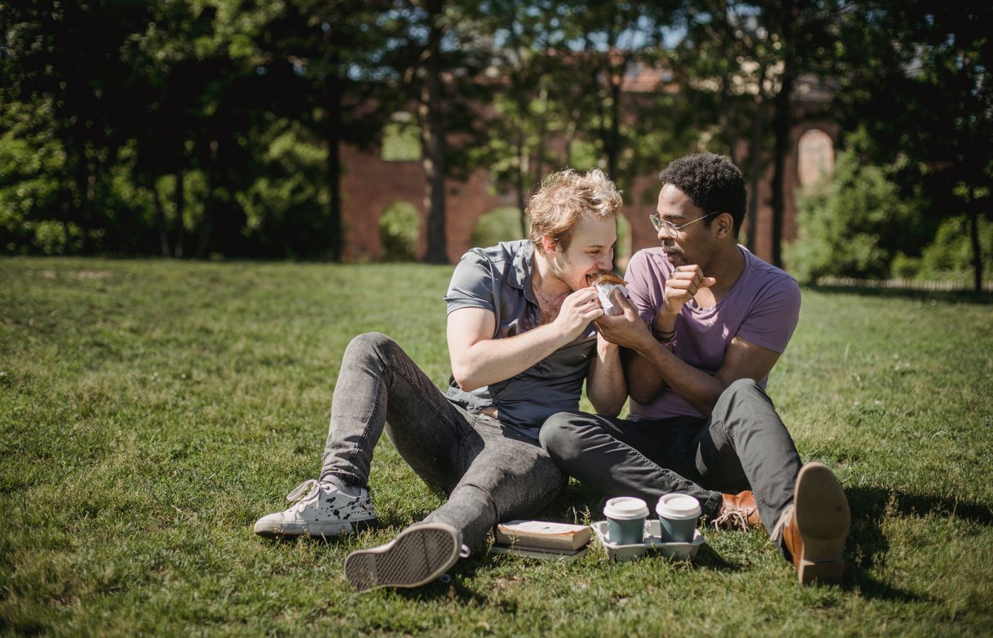 Interracial gay couple enjoys a sunny day in the park sharing a meal, expressing love and happiness.