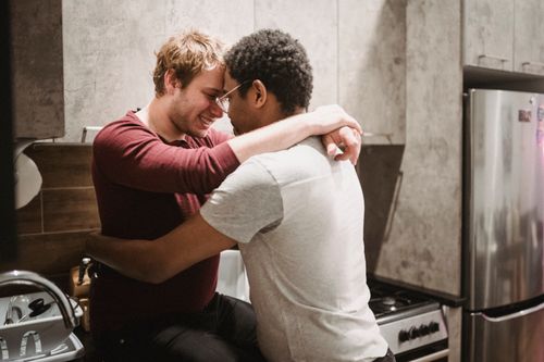 Free Interracial gay couple sharing a tender embrace in a cozy kitchen setting, smiling and expressing love. Stock Photo