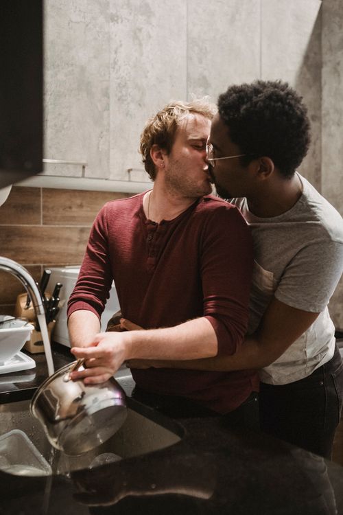 Free An interracial gay couple embraces and kisses while washing dishes in a cozy kitchen setting. Stock Photo