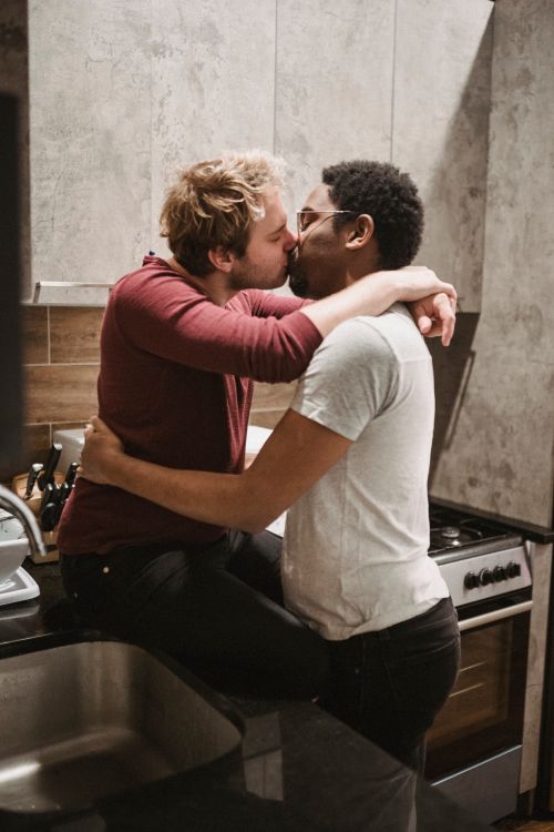 Free A loving interracial gay couple shares a tender kiss in a modern kitchen setting, symbolizing affection and togetherness. Stock Photo