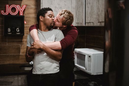 A loving interracial gay couple shares a kiss in their cozy kitchen, embracing affection and joy. — photo by Ketut Subiyanto