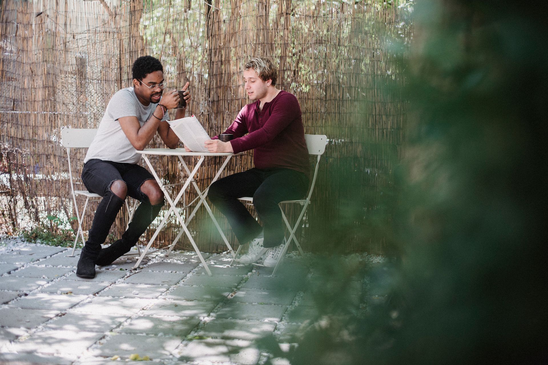 Two men enjoy a relaxed conversation outdoors over a newspaper at a patio table.