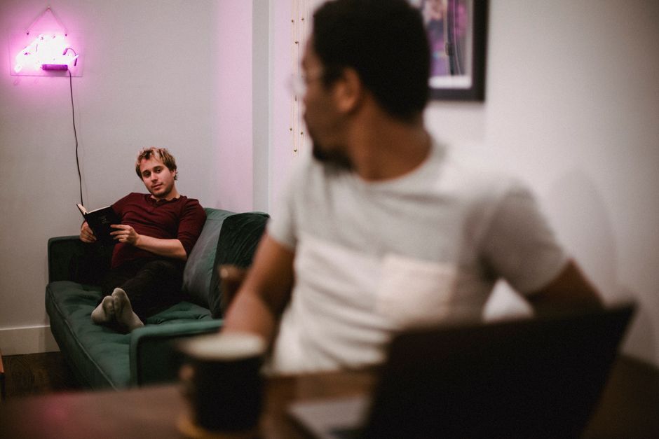 Two men relaxing indoors, one reading on a sofa, the other working on a laptop with coffee. Two men relaxing indoors, one reading on a sofa, the other working on a laptop with coffee.