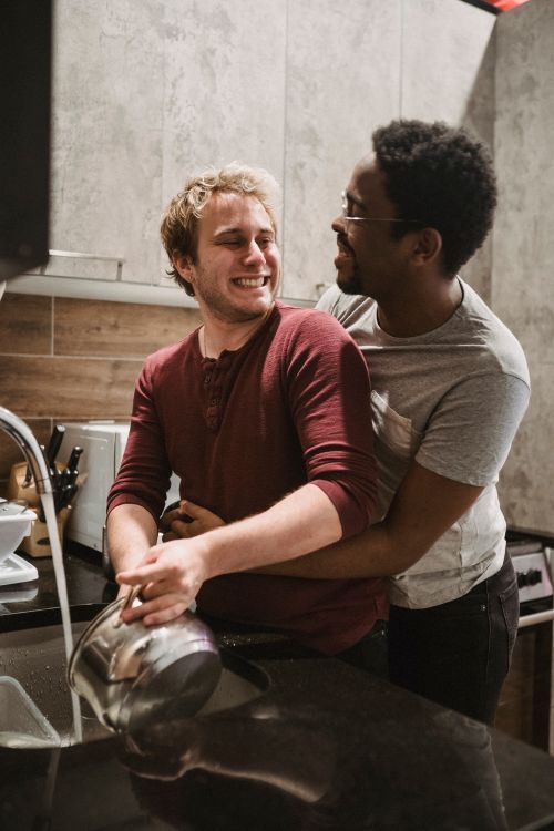 Free A joyful interracial couple in a kitchen sharing an affectionate embrace while washing dishes. Stock Photo