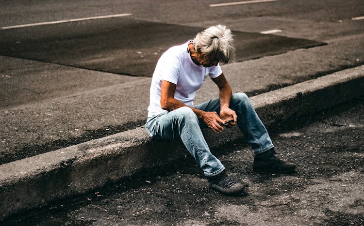 A Man Sitting On The Curb While Using His Phone